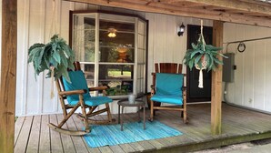 Eine hölzerne Veranda mit zwei Schaukelstühlen, einem kleinen Tisch, einem Fenster mit Vorhängen und einer hängenden Pflanze.