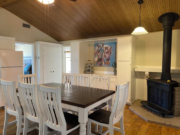 A dining area with a wooden table and white chairs, a painting of horses on the wall, and a black stove.
