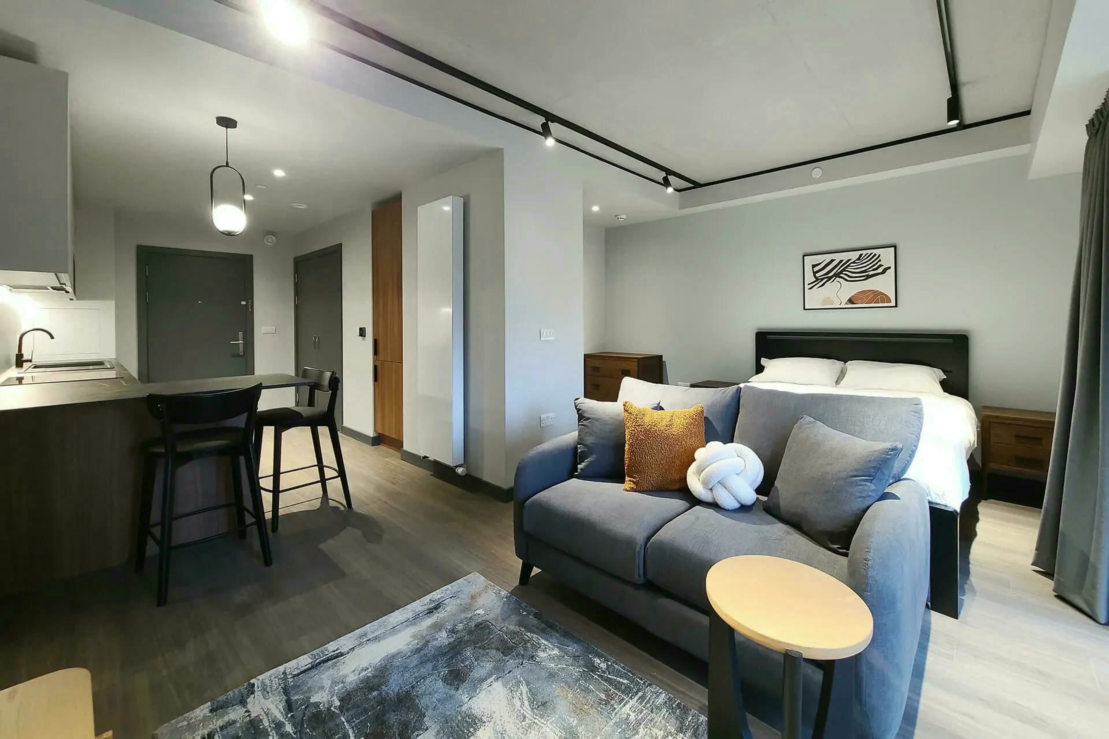 A modern hotel room with a sofa, a small round table, a bed, a desk, and a kitchen area.