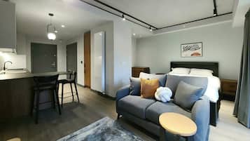 A modern hotel room with a sofa, a small round table, a bed, a desk, and a kitchen area.