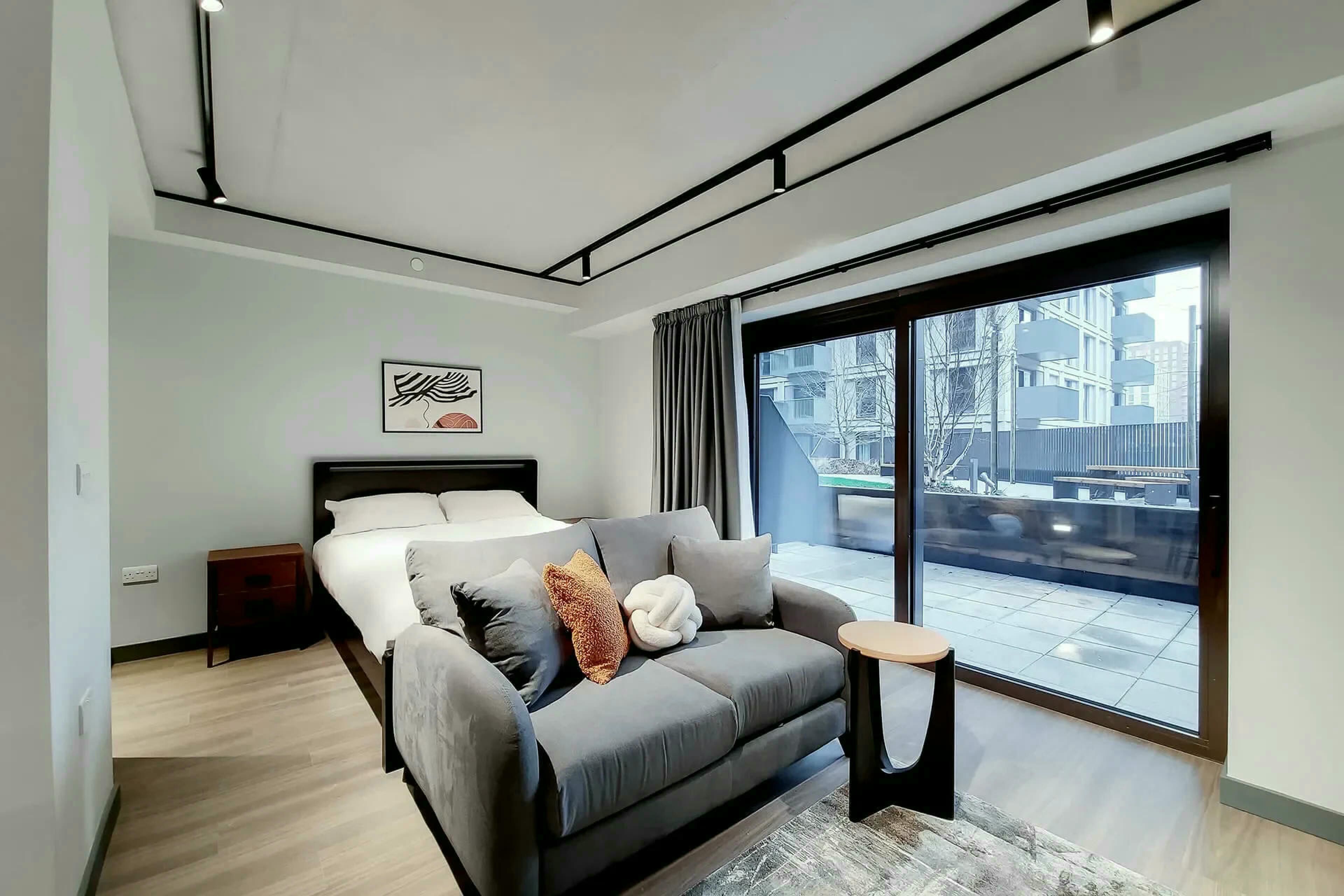 A modern bedroom with a bed, a grey sofa, a small table, and a large window offering a view of buildings.