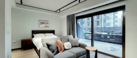 A modern bedroom with a bed, a grey sofa, a small table, and a large window offering a view of buildings.