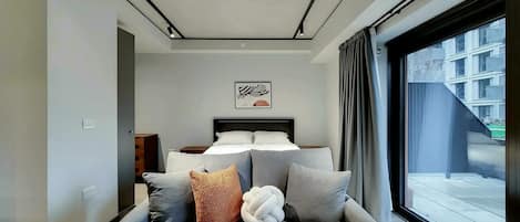 A modern hotel room with a bed, a sofa, and a large window offering a view of the city.