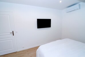 A room with a bed, a door, a wall-mounted TV, and an air conditioning unit. - Domaine de L'Olymp (Cornant)