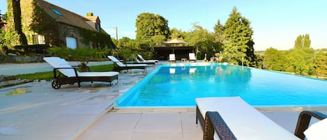 A rectangular swimming pool with lounge chairs and a covered area, surrounded by greenery and a building with a sloped roof.