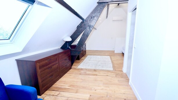 Una stanza con soffitto spiovente, pavimento in legno, un divano blu, un comò in legno scuro e un piccolo tappeto.