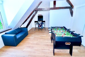 A room with a foosball table, a blue sofa, and a view of a balcony. - Domaine de L'Olymp (Cornant)