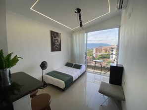 Ein modernes Schlafzimmer mit Bett, Schreibtisch, Stuhl, Ventilator und einem großen Fenster mit Blick auf die Stadt.