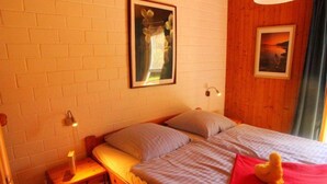 A single bed with a red pillow, a nightstand with a lamp, a framed picture on the wall, and a window with curtains. - Urlaub im Grunen - Doppelbungalow mit Terrasse im Ferienpark Mirow (Mirow)