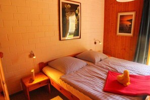 A single bed with a red pillow, a nightstand with a lamp, a framed picture on the wall, and a window with curtains. - Urlaub im Grunen - Doppelbungalow mit Terrasse im Ferienpark Mirow (Mirow)