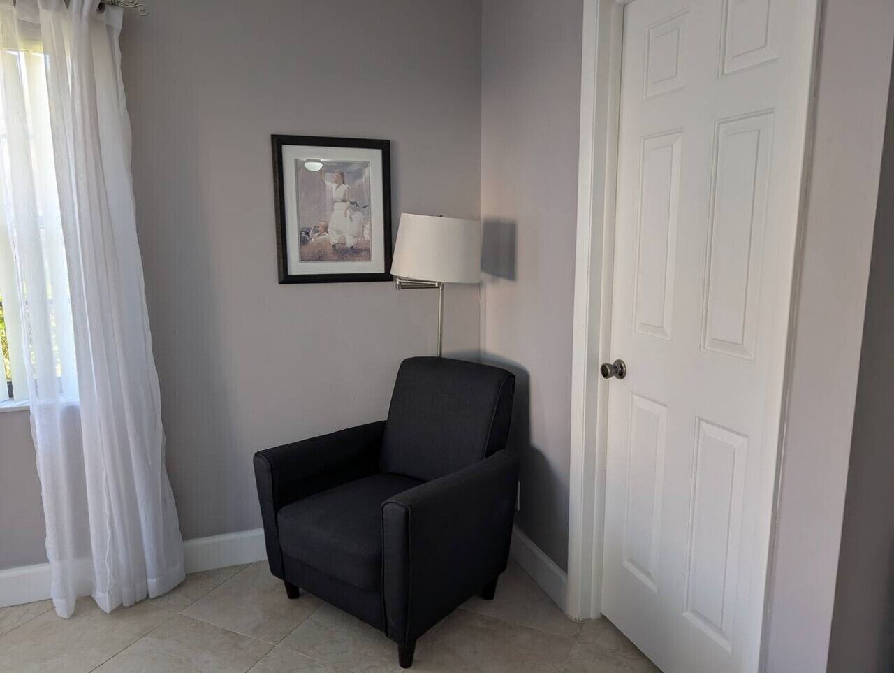 Une pièce avec un fauteuil noir, une porte blanche, un tableau encadré au mur et une lampe.