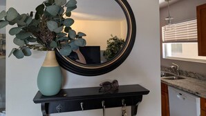Ein Badezimmer mit Spiegel, einer Vase mit grünen Blättern, einem schwarzen Regal mit einer Brille und einer Küchenzeile mit Spüle und Schränken.