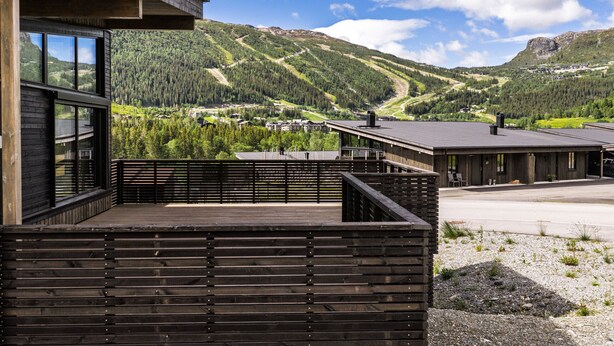 Un edificio moderno con terraza de madera, amplios ventanales y vistas a un paisaje montañoso.