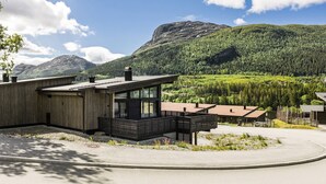 Un edificio moderno de madera con grandes ventanales, una terraza y vistas a una cadena montañosa boscosa.