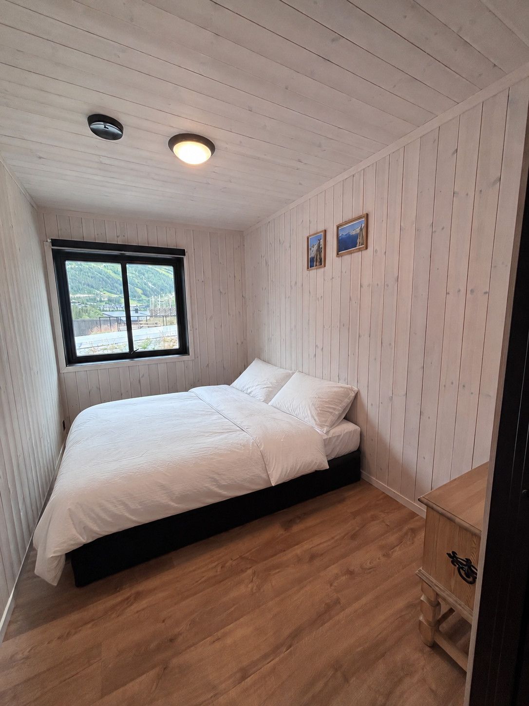 A bedroom with a bed, a window, wooden walls, and a wooden dresser.