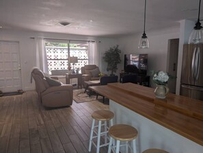 A modern kitchen with a wooden countertop and bar stools, a dining area with a sofa and chairs, and a large window with curtains. - Cozy house with heated pool near the beach (Lighthouse Point)