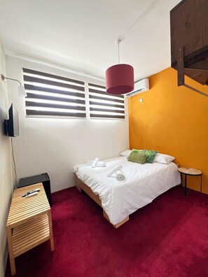 Ein Schlafzimmer mit einem Bett, einem kleinen Holztisch, einer roten Hängelampe und einem Fenster mit Jalousien.