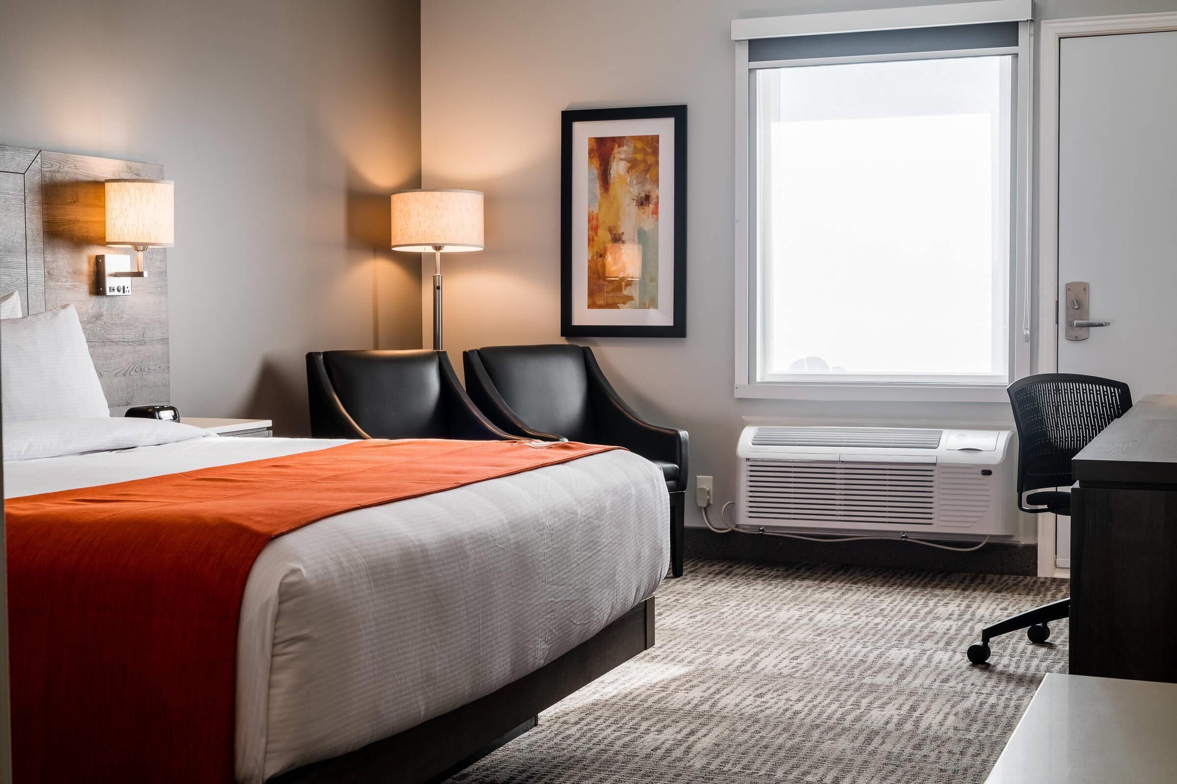 A hotel room with a bed, two chairs, a desk, a lamp, a painting, and a window.