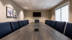 A conference room with a long table, blue upholstered chairs, a flat-screen TV on the wall, and a window on one side. - Amsterdam Inn & Suites, An Ascend Collection Hotel (Grand Falls)