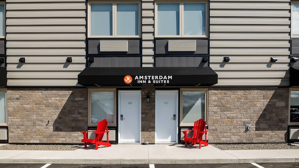 A two-story building with a sign for 'AMSTERDAM INN & SUITES' above the entrance. There are two red chairs outside the doors. - Amsterdam Inn & Suites, An Ascend Collection Hotel (Grand Falls)