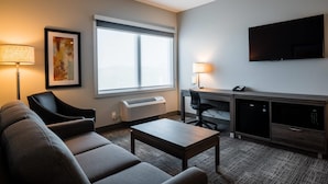 A hotel room with a sofa, a small table, a desk with a chair, a flat-screen TV, and a window with an air conditioner. - Amsterdam Inn & Suites, An Ascend Collection Hotel (Grand Falls)