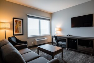 A hotel room with a sofa, a small table, a desk with a chair, a flat-screen TV, and a window with an air conditioner.