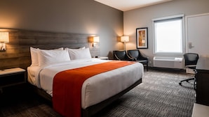 A hotel room with a large bed, bedside tables, a desk with a chair, a window with blinds, and a wall-mounted air conditioner. - Amsterdam Inn & Suites, An Ascend Collection Hotel (Grand Falls)