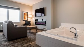A modern hotel room with a sofa, a flat-screen TV, a large bathtub, and a small table with two wine glasses.