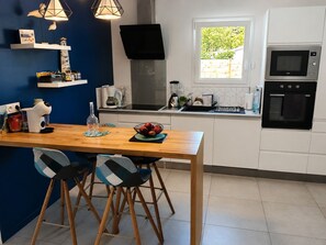 A modern kitchen with white cabinets, a wooden dining table, two bar stools, a built-in oven, a microwave, and a window with a view of greenery. - Le Hâvre des Dunes - Piscine (Brem-sur-Mer)