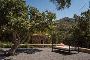 A bed with a metal frame and a cushion, set on a gravel surface, surrounded by trees and a stone wall, with a mountainous landscape in the background.