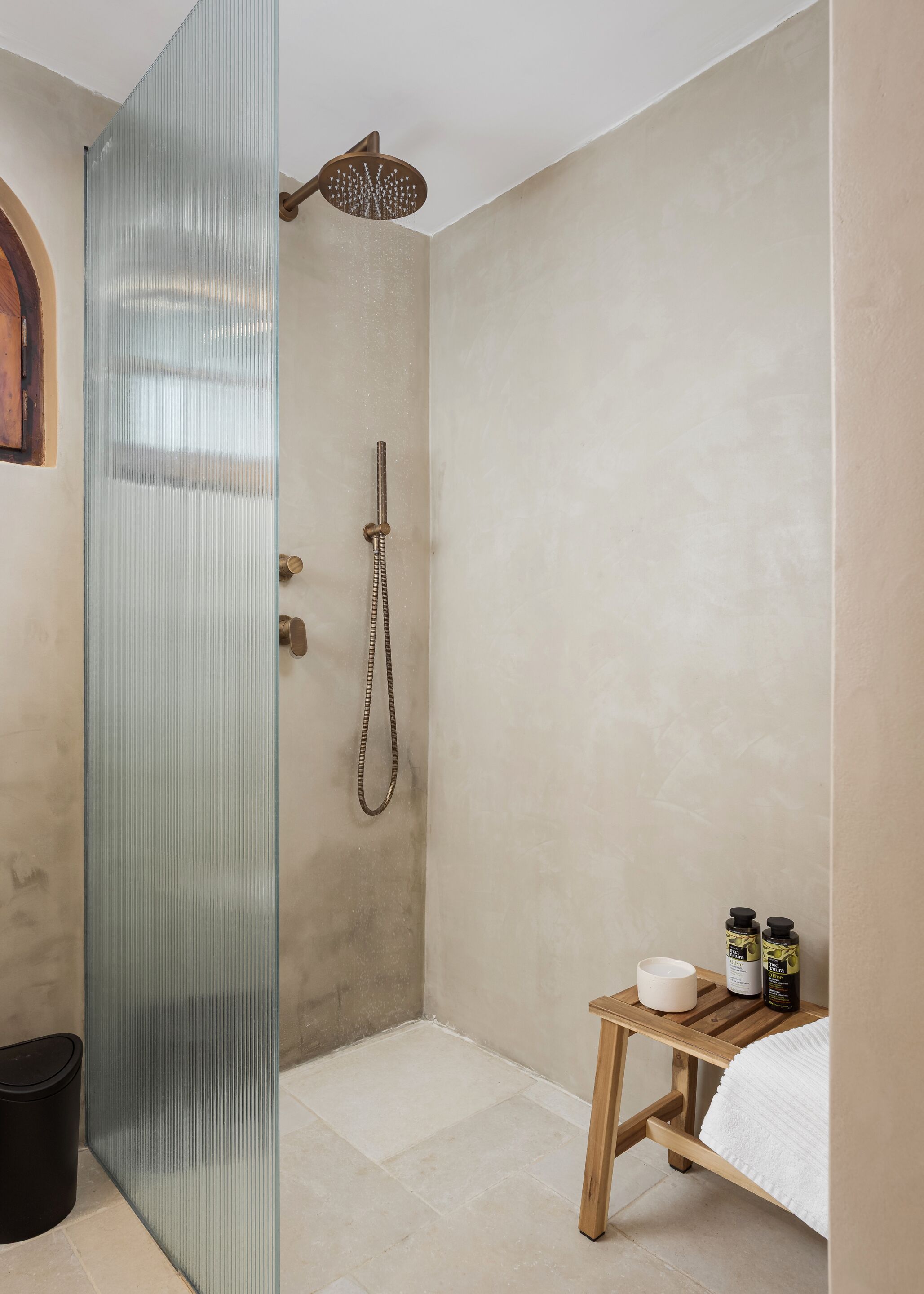 Un baño con mampara de vidrio, ducha con cabezal de ducha manual, banca de madera con artículos de aseo personal y un cesto de basura.