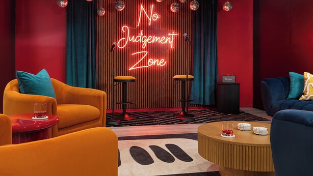 Una habitación con un letrero de neón que dice 'No Judgement Zone', dos taburetes de bar, una mesa roja con un vaso y un sofá azul con un cojín.
