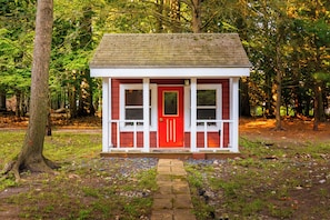 Ein kleines, eingeschossiges Holzhaus mit roter Tür und weißer Verzierung, umgeben von Bäumen.