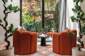 Une pièce avec une grande fenêtre, une table ronde sur laquelle se trouve une théière, et deux chaises orange.