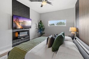Un dormitorio moderno con un televisor de pantalla plana en la pared, una cama con almohadas, una mesita de noche con lámpara y una ventana con persianas.