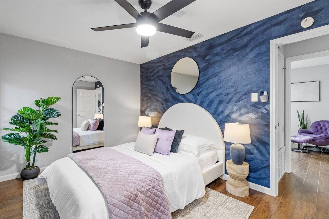 A bedroom with a bed, bedside lamps, a ceiling fan, a mirror, and a potted plant.