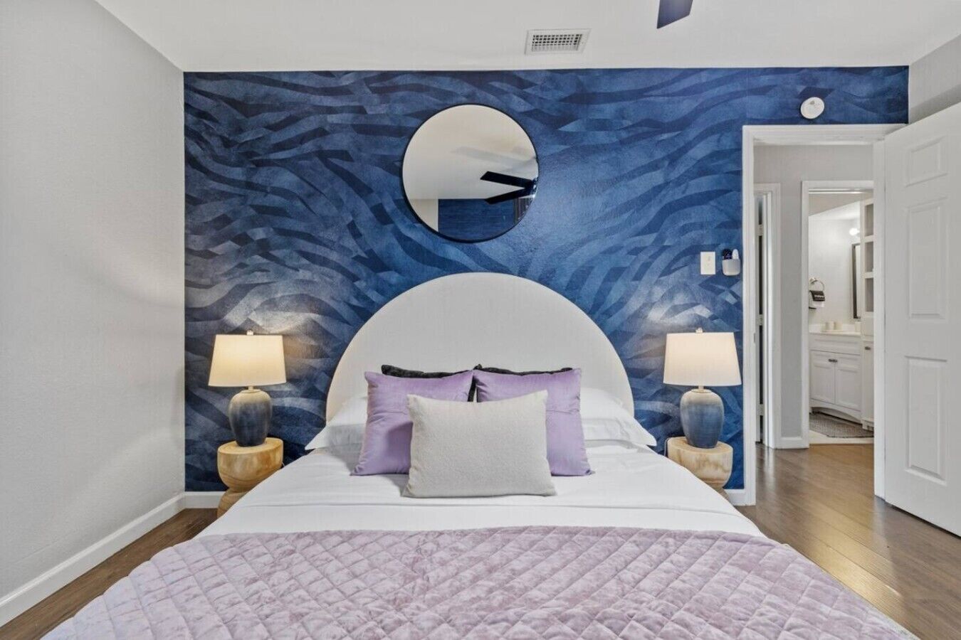 Une chambre à coucher avec un grand lit, deux tables de chevet avec des lampes, un grand miroir rond au-dessus du lit et un mur orné d’un motif bleu.