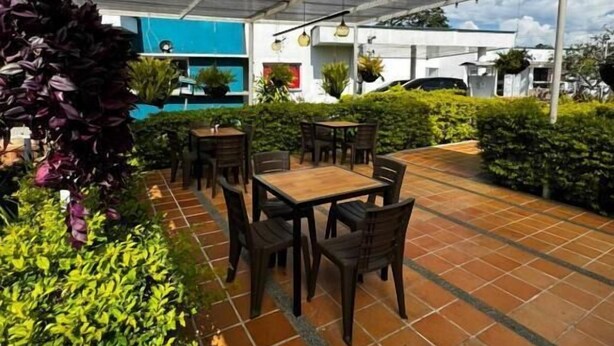 En udendørs terrasse med borde og stole, omgivet af grøn vegetation og et overdækket område med et metal tag.