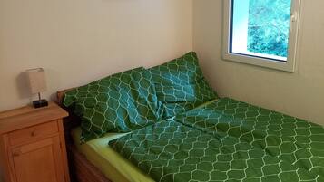 Ein Zimmer mit Holzboden, einem grün gemusterten Bett, einem kleinen Nachttisch aus Holz, einem gerahmten Bild an der Wand und einem Fenster mit Blick auf Bäume.