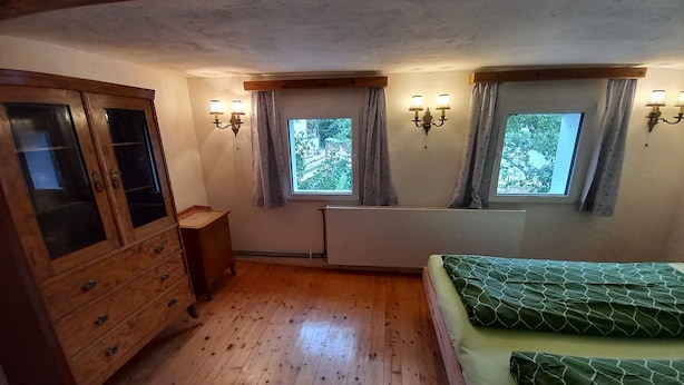 Ein Zimmer mit Holzboden, einem Bett mit grüner Bettwäsche, einem Holzschrank und zwei mit Vorhängen versehenen Fenstern.
