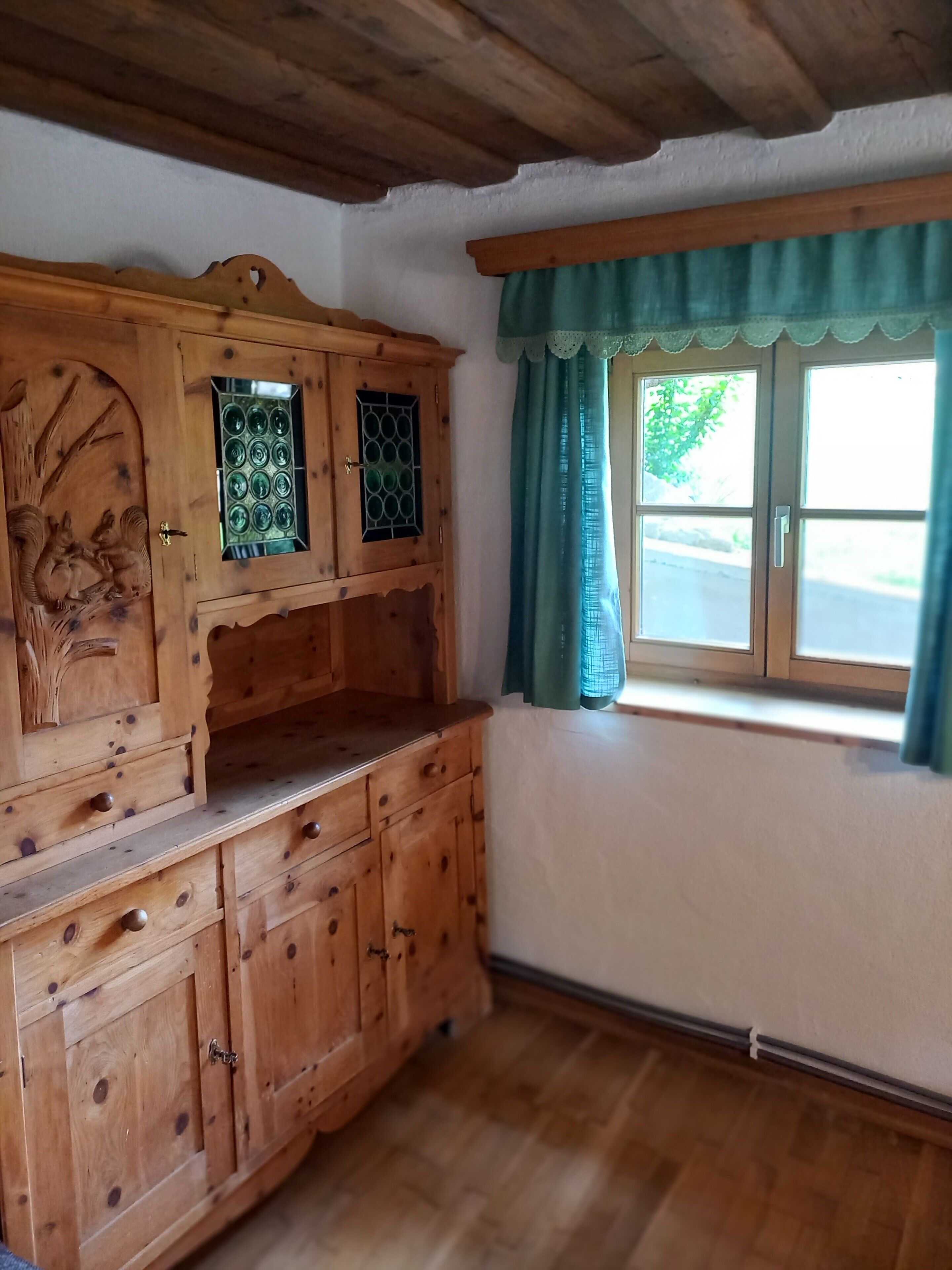 Ein Holzkabinett mit aufwendigen Schnitzereien, ein Fenster mit grünem Vorhang und ein Holzboden.