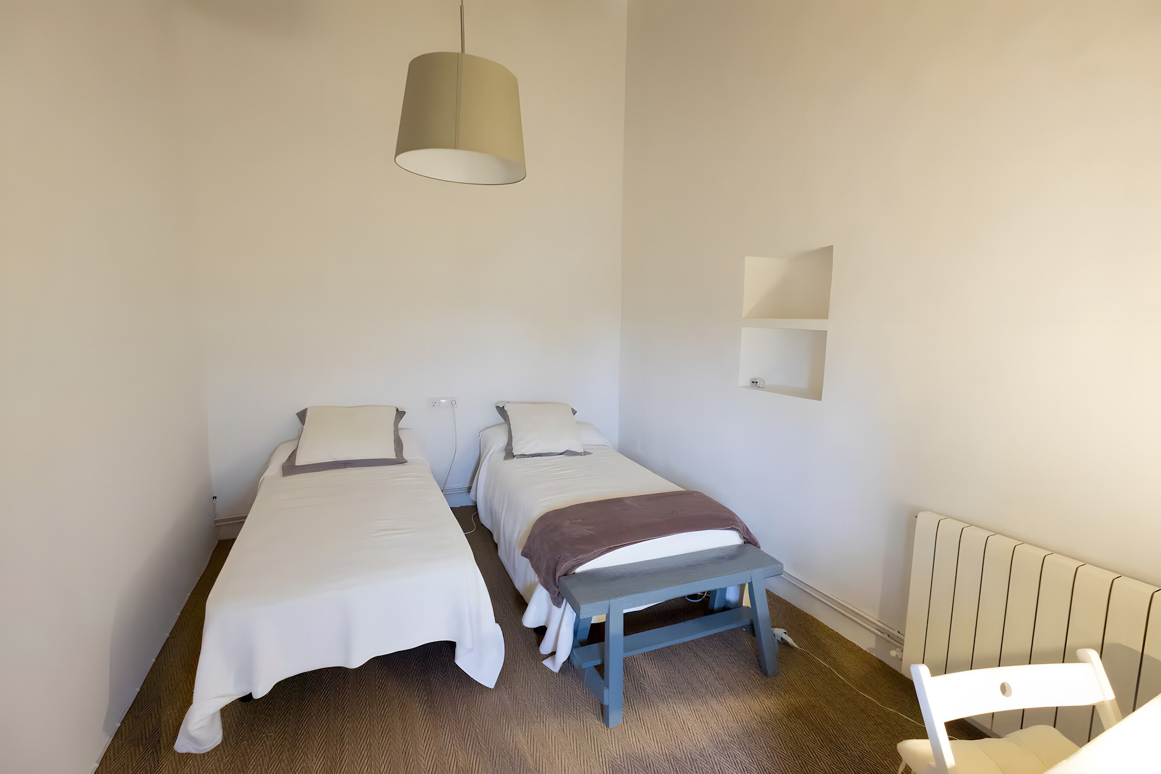 Una habitación sencilla y luminosa con dos camas individuales, un pequeño banco, una lámpara colgante y un radiador.