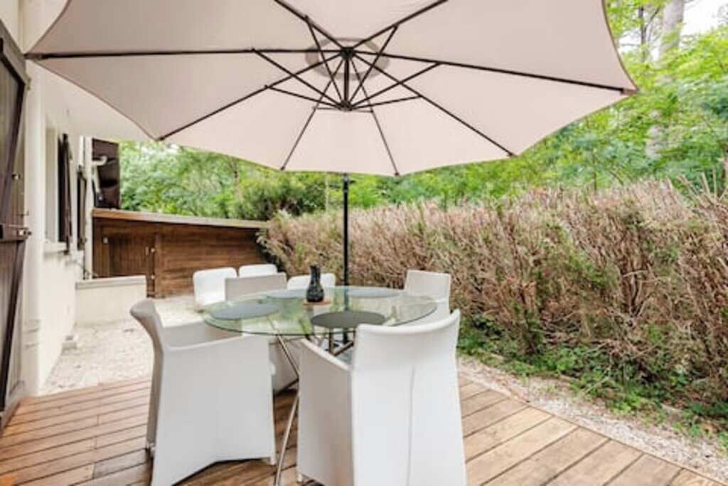 En udendørs terrasse med et glasbord og hvide stole, omgivet af et træterrasse og en stor parasol.