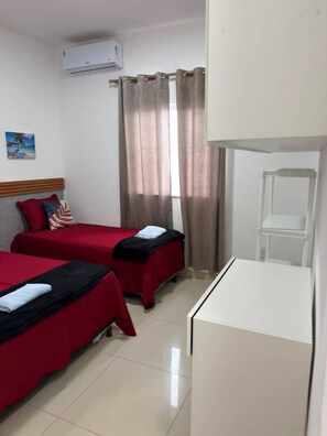 A hotel room with two beds, a white desk, an air conditioning unit, a window with curtains, and a small shelf.