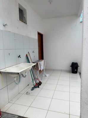 A room with a white tiled floor, a sink with a hose, a broom, and a trash bin.