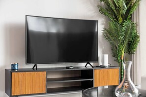 A modern living room with a flat-screen TV on a wooden stand, a small table with a remote, and a potted plant. - StayShort - Chic 1-Bedroom Apartment in Dubai Marina That Sleeps 2 (Dubai)