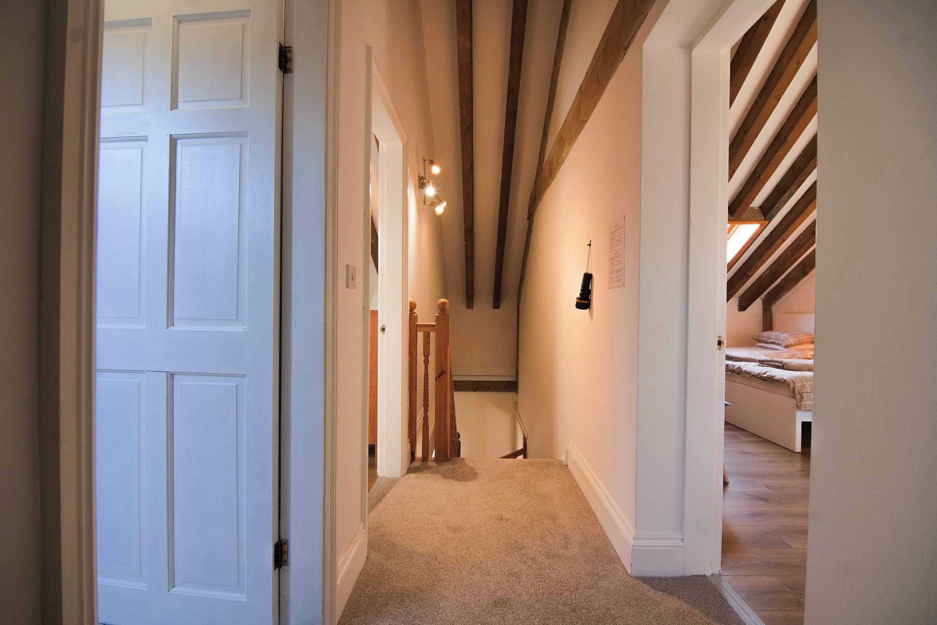 A hallway with a carpeted floor, wooden beams on the ceiling, and a bed in the adjacent room.