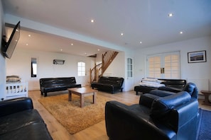 A spacious living room with a wooden staircase, black leather sofas, a wooden coffee table, and a flat-screen TV mounted on the wall.