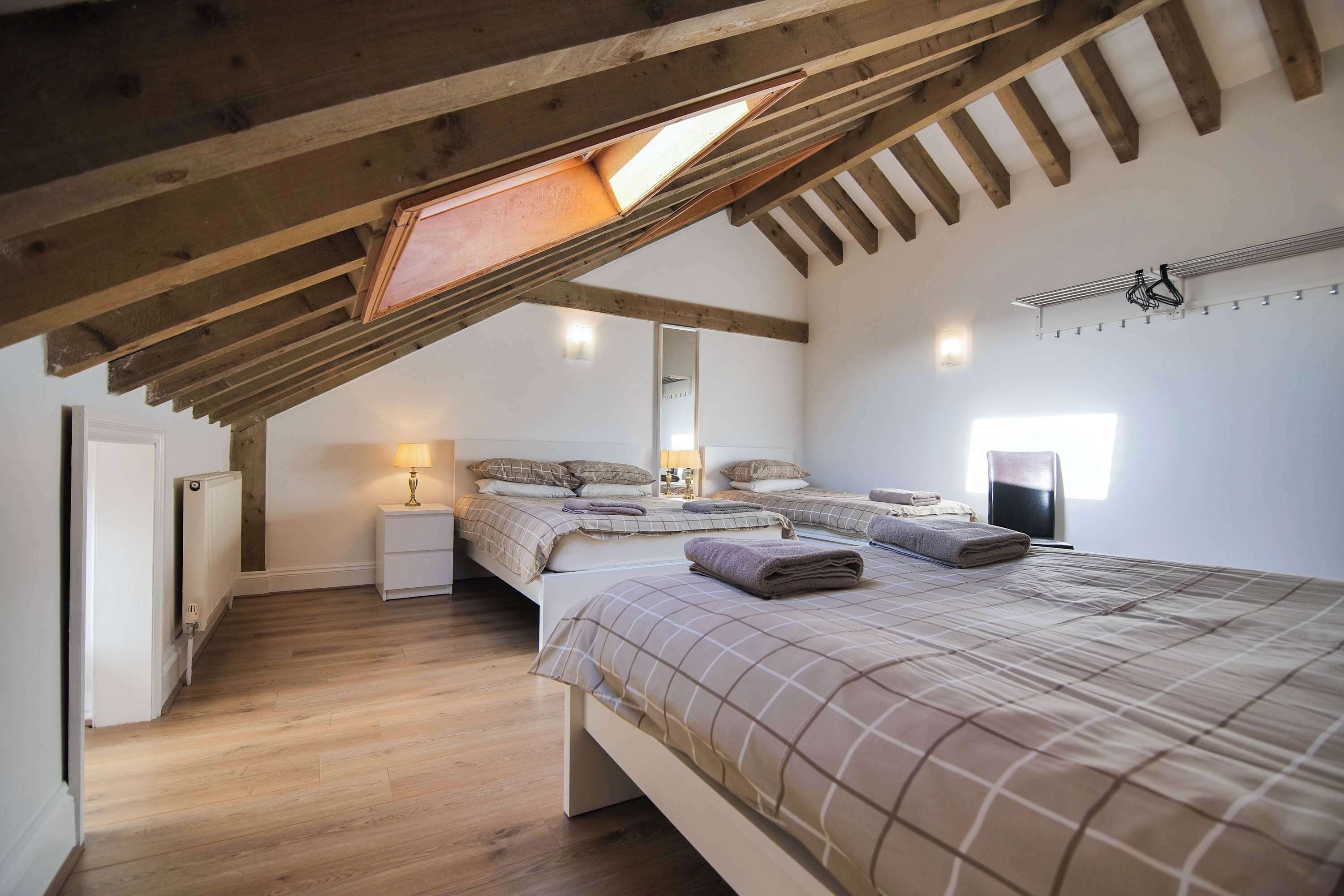 A bedroom with a sloped ceiling, wooden beams, a radiator, a bedside table with a lamp, two beds with towels, and a wall-mounted heater.