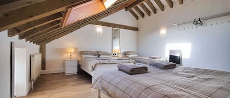A bedroom with a sloped ceiling, wooden beams, a radiator, a bedside table with a lamp, two beds with towels, and a wall-mounted heater.
